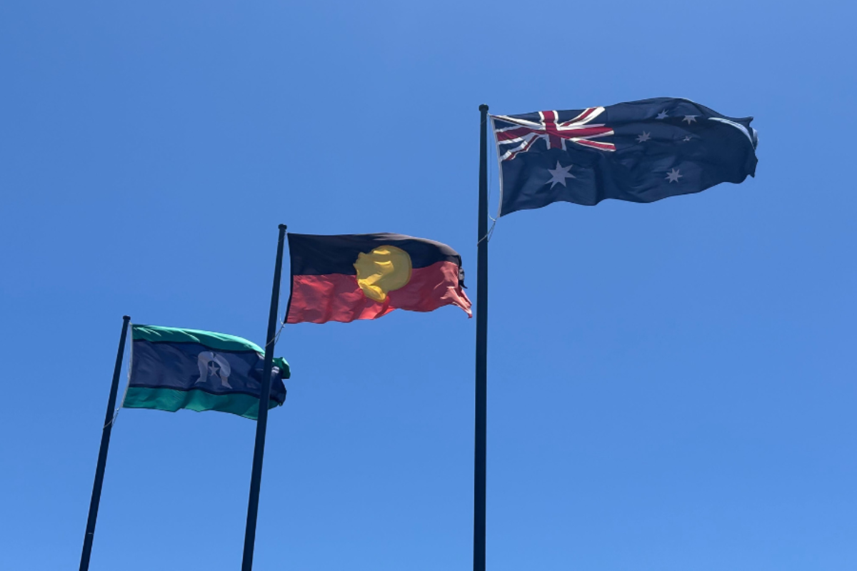 Three flags flying against a clear blue sky: the Torres Strait Islander flag, the Aboriginal flag, and the Australian national flag